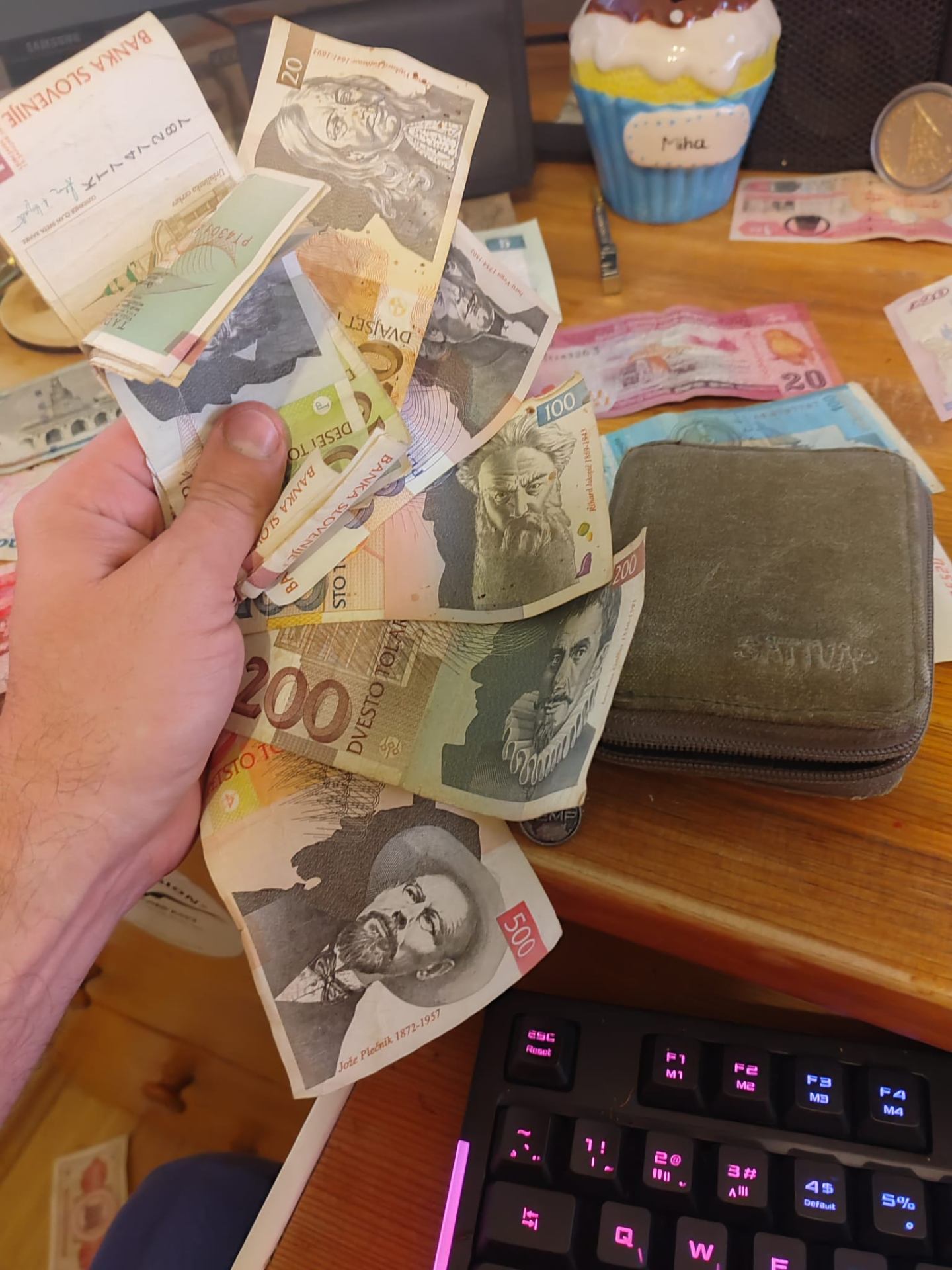 A hand holding a stack of Slovenian Tolar banknotes (10, 20, 100, 200, 500) featuring Plečnik, Adamič, Vega and Prešeren, pulled from a green hemp wallet on a wooden desk.